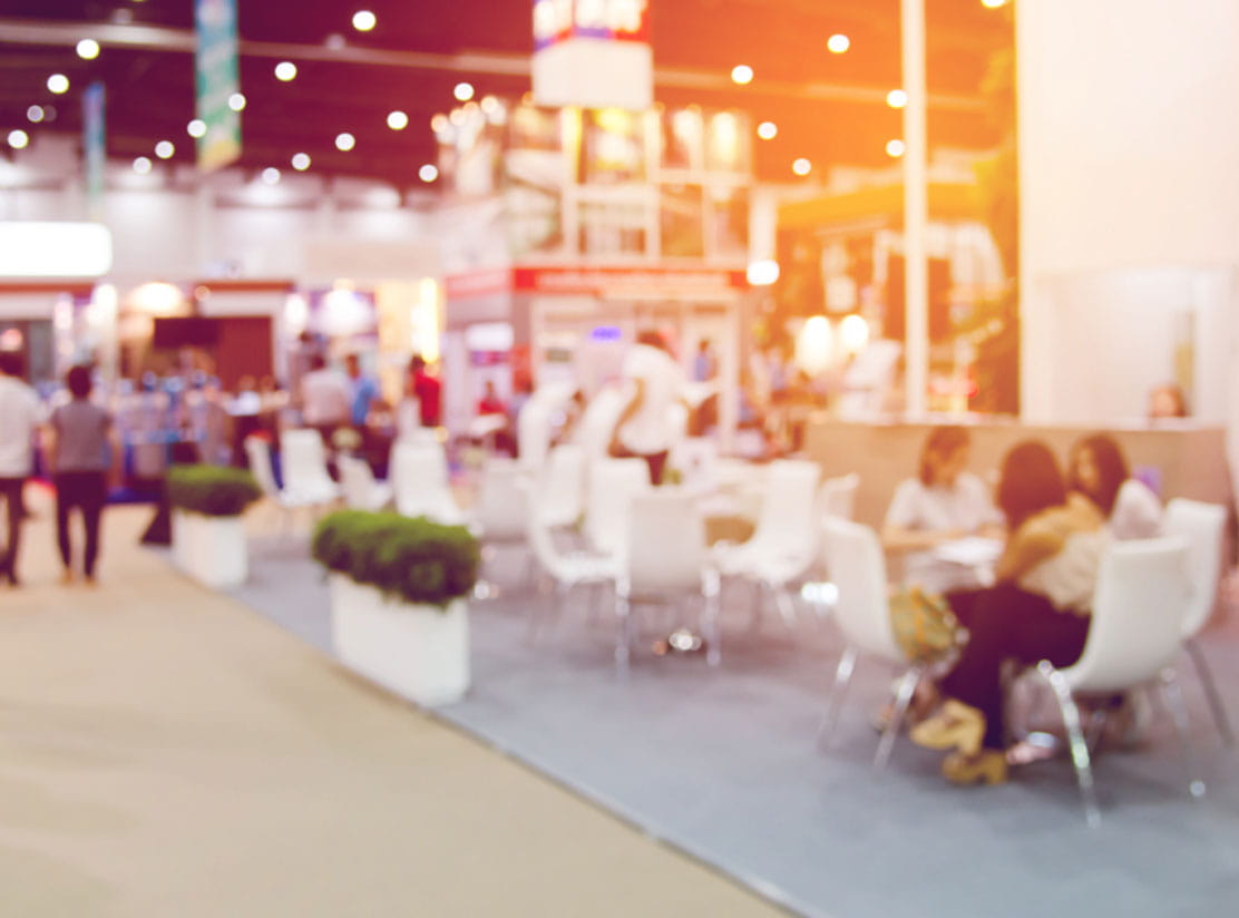 An indoor event space filled with people, featuring exhibition booths in the background.