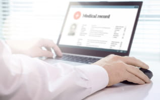 Close-up of a person in a white coat using a laptop displaying a medical record, with hands on the keyboard and mouse, in a brightly lit office.