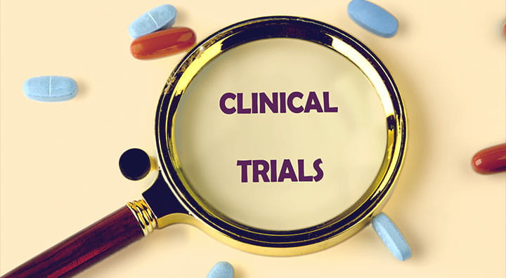 A magnifying glass focuses on the words "CLINICAL TRIALS," surrounded by colorful pills and a syringe on a beige background.