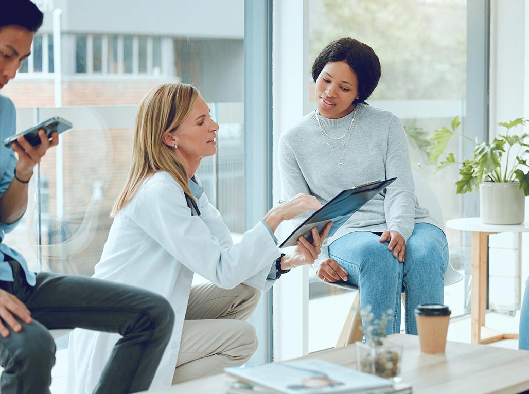 Doctor in white coat discusses medical information with a seated patient in a clinic waiting area.